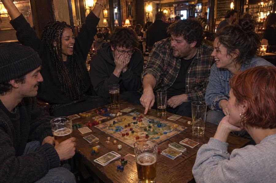 Six personnes jouent à un jeu de société dans un bar, une joueuse lève les bras de joie